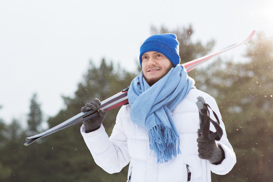 Young sportsman in white winter jacket, blue scarf and beanie hat holding pair of skis while spending time in winter park