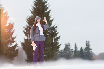Fototapeta na wymiar Pretty skier in winter activewear making selfie on smartphone in the middle of forest