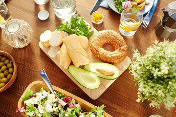 Fresh bread and avocado halves on wooden board surrounded by salad, olives, lemonade, candle and...