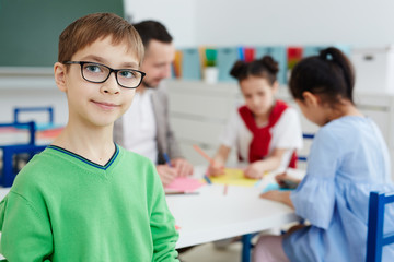 Young boy in eyeglasses looking at camer aon background of his classmates and teacher drawing