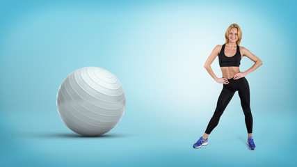 Fototapeta na wymiar A young fit blonde woman stands with hands on her hips near a large silver exercise ball.