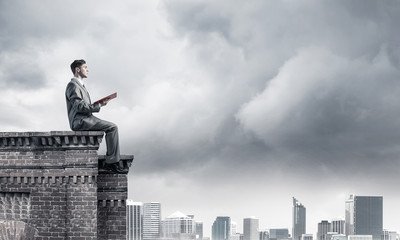 Man on roof edge reading book and cityscape at background