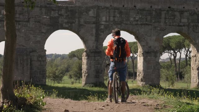 Cyclist with bike reads map for direction in front of ancient roman aqueduct in a park. Young attractive athletic man with orange sportswear and backpack in parco degli acquedotti in Rome under a tree