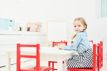 Cute little girl looking at camera while sitting by table on red chair in kindergarten