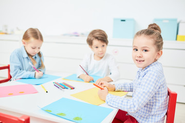 Three creative kids with crayons drawing by table in kindergarten