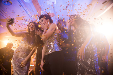 Group  of beautiful young women wearing glittering dresses dancing under golden confetti and taking selfies enjoying raving party in nightclub - obrazy, fototapety, plakaty