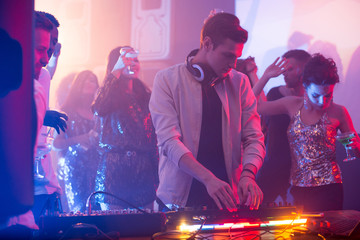 Portrait of handsome young DJ standing at mixer making music during night party in club, scene lit by stage lights with beautiful girls wearing glittering tops dancing in background