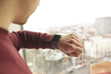 The man stands and is looking on his smart watches on his hand in front of window with view on city