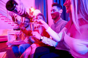 Side view at group of glamorous  people enjoying private party, sitting on sofa laughing happily while waitress pouring drinks