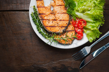 Grilled salmon steak served with grilled tomatoes, salad, lemon sliced, pepper seeds, rosemary, parsley on white plate on wooden table.