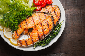 Grilled salmon steak served with grilled tomatoes, salad, lemon sliced, pepper seeds, rosemary, parsley on white plate on wooden table.