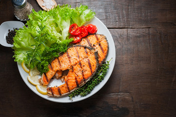 Grilled salmon steak served with grilled tomatoes, salad, lemon sliced, pepper seeds, rosemary, parsley on white plate on wooden table.