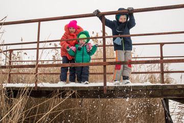 Cute little girl and boys in colorful winter clothes, playing on the bridge over a frozen river, outdoors during a snowfall. Active recreation in nature with children in winter