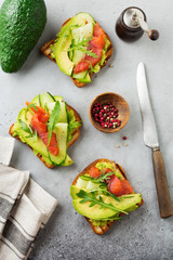 Open toast with trout, salmon, avocado, cucumber and arugula on wooden stand on gray concrete background. Selective focus. Top view. Copy space
