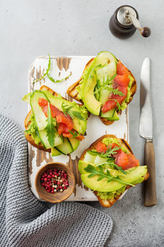 Open toast with trout, salmon, avocado, cucumber and arugula on wooden stand on gray concrete background. Selective focus. Top view. Copy space