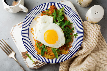 Fried egg with potato pancake, arugula and avocado on ceramic plate for breakfast on gray old concrete background. Selective focus. Top view. Copy space.