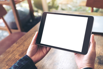 Mockup image of hands holding black tablet pc with blank desktop white screen on wooden table in cafe background