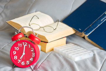 Red alarm clock with open book on strack and pills box on bed