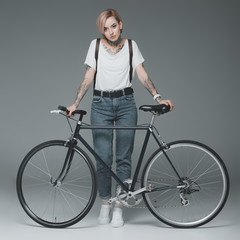 Obraz premium beautiful stylish girl with tattoos standing with bicycle and looking at camera isolated on grey