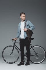 full length view of young tattooed man standing with bicycle and looking away on grey