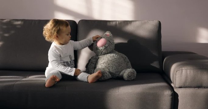 Girl with light hair play with grey toy cat on a stylish couch in sunny day