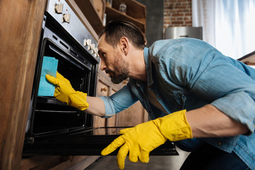 Clean oven. Appealing bearded male cleaner leaning over oven while cleaning it and wearing gloves