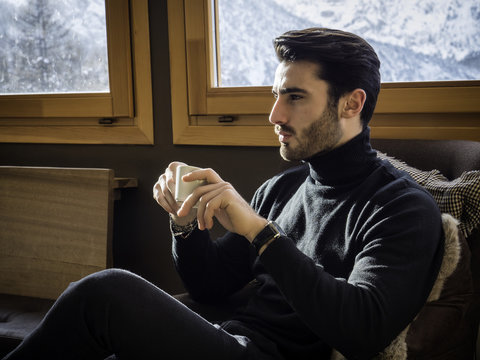 Young handsome man with beard drinking coffee or tea or hot chocolate, looking outside the window, away confidently on background of snowy mountains.