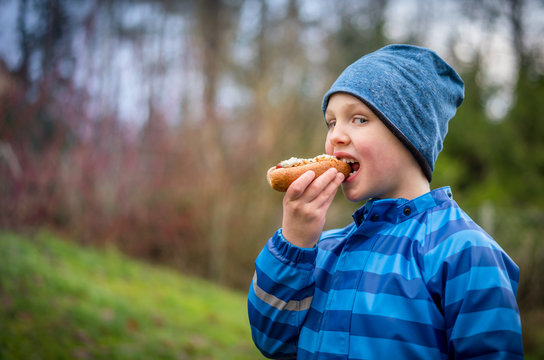 Boy in blue winter rain jacket and hat eating hotdog outdoors, profile view.
