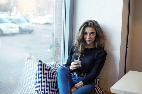 Indoor shot of fashionable young woman in trendy clothes spending morning at cafeteria browsing social media on smart phone, shopping online, sitting on windowsill, having happy facial expression