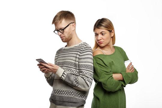 Studio shot of suspicious distrustful young blonde female in green top standing next to her bearded boyfriend, looking over his shoulder, spying over him while he is typing message using mobile phone