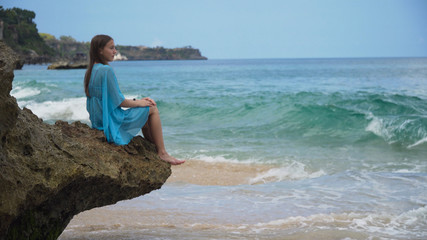 Beautiful girl in blue dress sitting on a rock above the sea and looking at ocean. Travel concept.