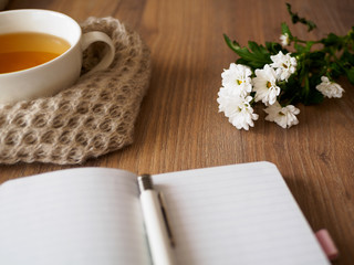 Eine Tasse Tee, Wollschal, Notizbuch, Kugelschreiber und weiße Blumen auf einem braunen Holztisch