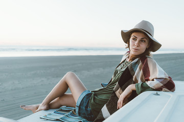 Charming young girl with hat, poncho, backpack with nice smile sitting on roof of recreational vehicle and relaxing on the beach at sunset in warm weather. Boohoo style.