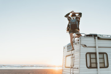 Charming woman with hat, poncho,  backpack standing on roof of recreational vehicle on the ocean...