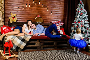 Portrait of happy family with little beautiful daughter resting on the wooden sofa with warm red blanket against wooden wall, near little girl. Concept of celebration and New Year