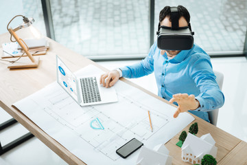 Concentrated on work. The top view of a pleasant young man working on the laptop and using a VR headset, visualizing the constructions