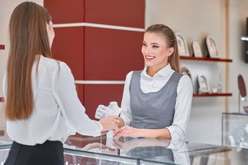 Pretty young female jewelry worker in white blouse and grey uniform is helping to choose a necklace for a female visitor wearing smart clothes such as black skirt and white blouse in a jewelry store.