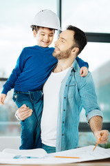 Best duo. Happy young architect carrying his little son in a hard hat while working on a blueprint...