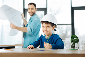 Future artist. Pleasant young architect holding a blueprint and looking at his son with affection...