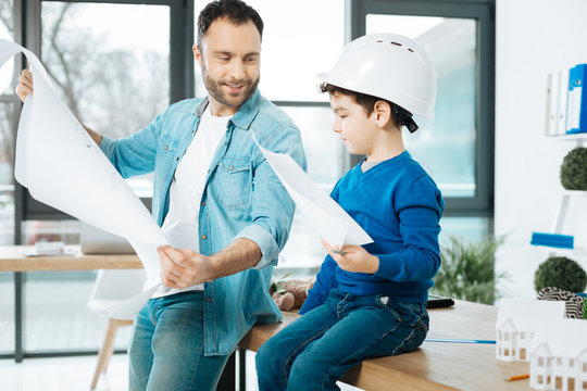 Skilled artists. Cute little boy in a white hard hat holding a picture while sitting on the table next to his father showing a blueprint to him