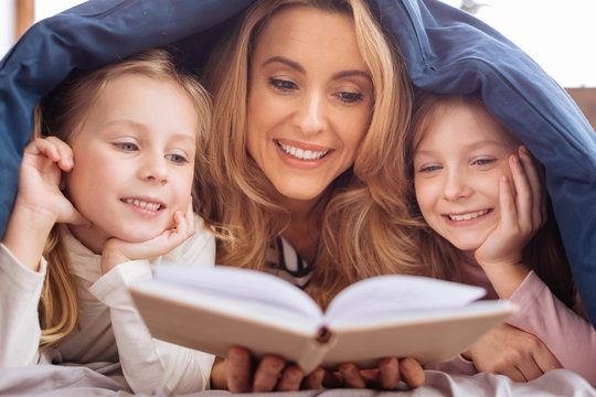 Reading. Devoted content blond mother and her little daughters smiling and reading a book while lying on the couch
