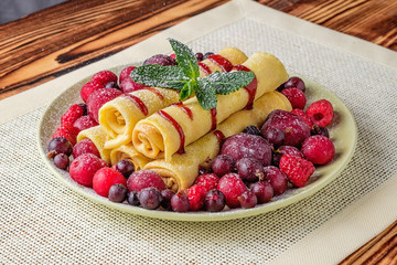 On the table is a white plate with pile of rolled pancakes. Pancakes are poured with jam and decorated with green mint leaves and around them are berries, sprinkled with powdered sugar.