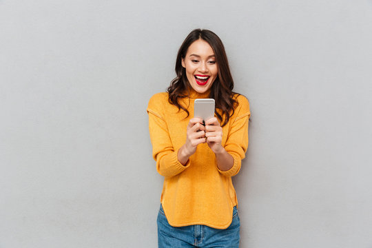 Image of Surprised happy brunette woman in sweater using smartphone