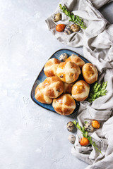 Homemade Easter traditional hot cross buns on blue plate with textile and colored quail eggs over white texture background. Top view, copy space