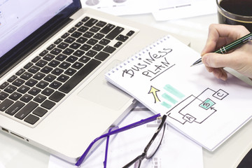 Business, Finance, Analysis concept. A workplace with a laptop. Woman's hands writing business plan in a notebook, light background