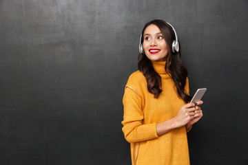 Smiling woman in sweater and headphones listening music by smartphone