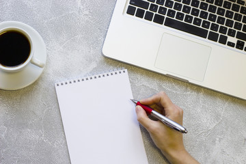 Modern office table desk, flat lay, top view. Workplace with a laptop, coffee cup, woman is redy to write in a blank notepad, grey concrete background