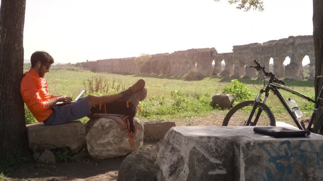 Cyclist with bike sitting on a rock under a tree writing on laptop in front of ancient roman aqueduct. Young attractive athletic man with orange sportswear and backpack in parco degli acquedotti in
