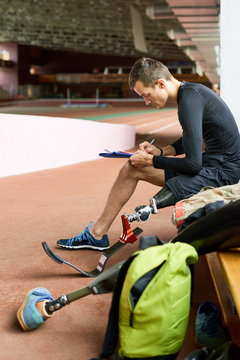 Side view portrait of young disabled athlete relaxing on bench in gym and  writing something, copy space