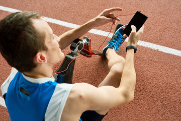 High angle view of young amputee athlete listening to music from smartphone  sitting on running track in modern stadium